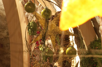 Der Fokus liegt auf dem Hintergrund, einem hängenden Weihnachtsgesteck, geschmückt mit goldenen Sternen, grünen Christbaumkugeln und grünen, glitzernden Elefanten, alles mit Draht auf einen Holzrahmen gespannt. Davor ist unscharf ein Teil eines gelb leuchtenden Sternes und weiterer Draht zu sehen.