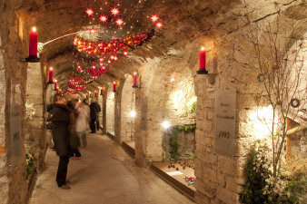 Ein Gang in einem Keller, geschmückt mit roten Kerzen an den Säulen zwischen den beleuchteten Nieschen und einem großen Schmuckband aus Draht entlang der Decke mit roten Weihnachtskugeln und Lichtern verziert.