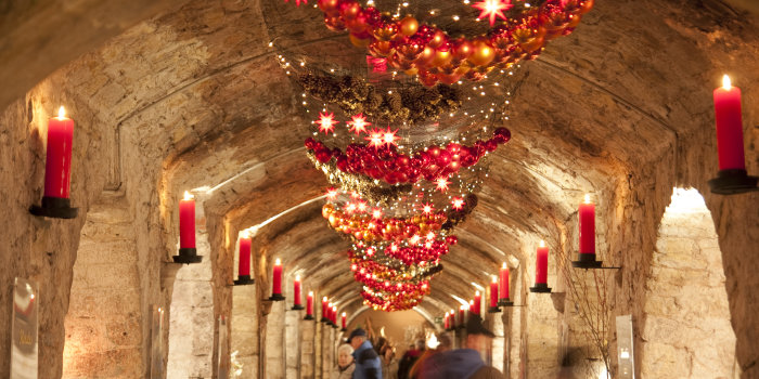 Der Gang eines Gewölbekellers, seitlich geschmückt mit roten Kerzen, im Fokus ein Schmuckband aus Draht, welches mit roten Weihnachtskugeln, Tannenzapfen und roten Lichtern in Sternenform verziert ist.
