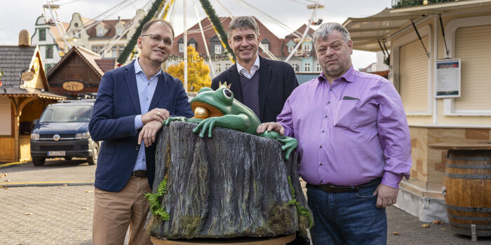drei Personen stehen an einer Froschfigur, die auf einem künstlichen Baumstamm sitzt