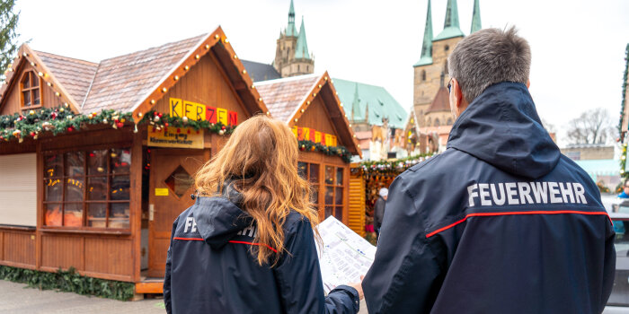 Eine Frau und ein Mann in Rückansicht. Beide tragen eine Jacke mit der Aufschrift „Feuerwehr“. 