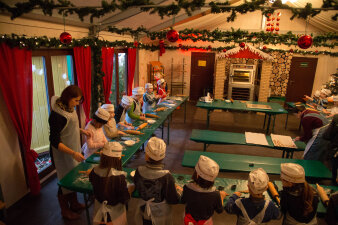 Kinder stehen mit Schürze und Mütze bekleidet in einer Reihe an langen Tischen und bearbeiten mit Unterstützung der Bäckerinnen den Plätzchenteig.
